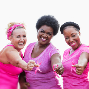 Three women smile while holding up pink ribbons and wearing pink to signify Breast Cancer Awareness Month