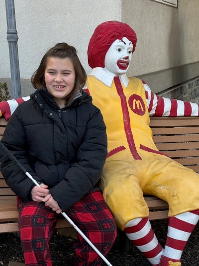 Young brunette girl with mobility cane site next to Ronald McDonald statue on bench
