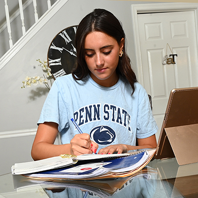 Teenage girl in blue Penn State t-shirt writing in notebook with clock and door in background