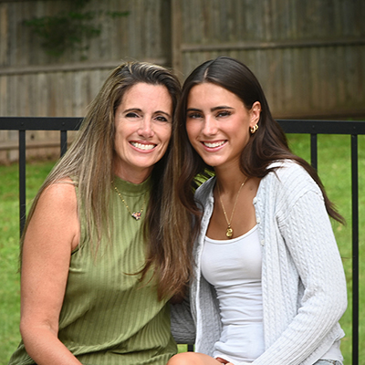 Smiling woman in green shirt posing with smiling young girl in white shirt with grassy backyard in background