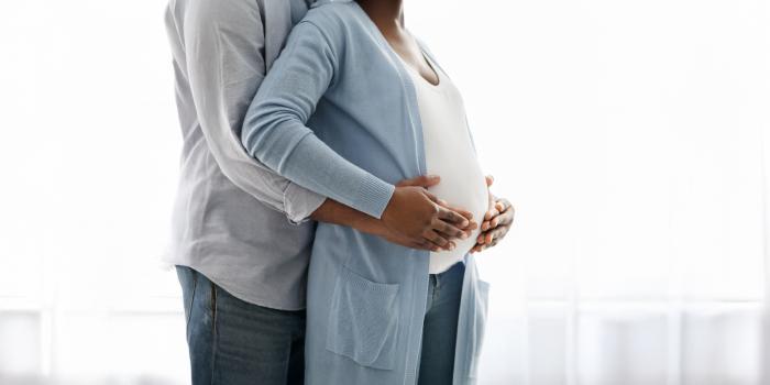 A pregnant woman and a man stand together in front of a large window, sharing a moment of anticipation.